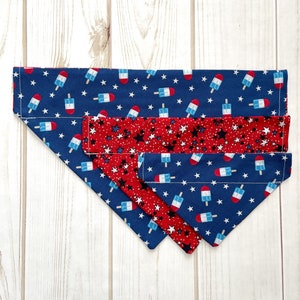 May include: Three layered dog bandanas in blue and red with a white star and popsicle pattern. The top layer is blue with white stars and red, white, and blue popsicles. The middle layer is red with white stars and black stars. The bottom layer is blue with white stars and red, white, and blue popsicles.