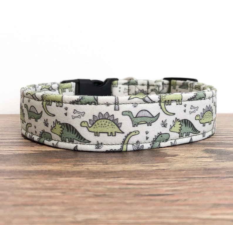 May include: A white dog collar with a green and gray dinosaur print. The collar has a black plastic buckle.