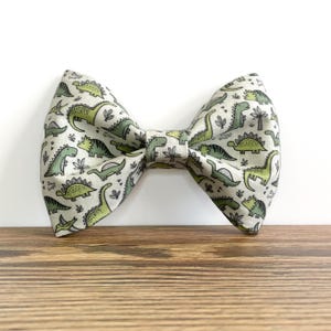 May include: A bow tie featuring a repeating pattern of various green dinosaurs and small black botanical illustrations on a light beige background. The fabric has a structured, folded shape with a central knot detail and a classic bow tie silhouette.