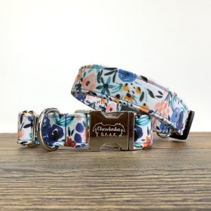May include: A white dog collar with a floral pattern in shades of blue, pink, orange, and green. The collar has a silver buckle with the engraved text "Chewlinka G.E.A.K."