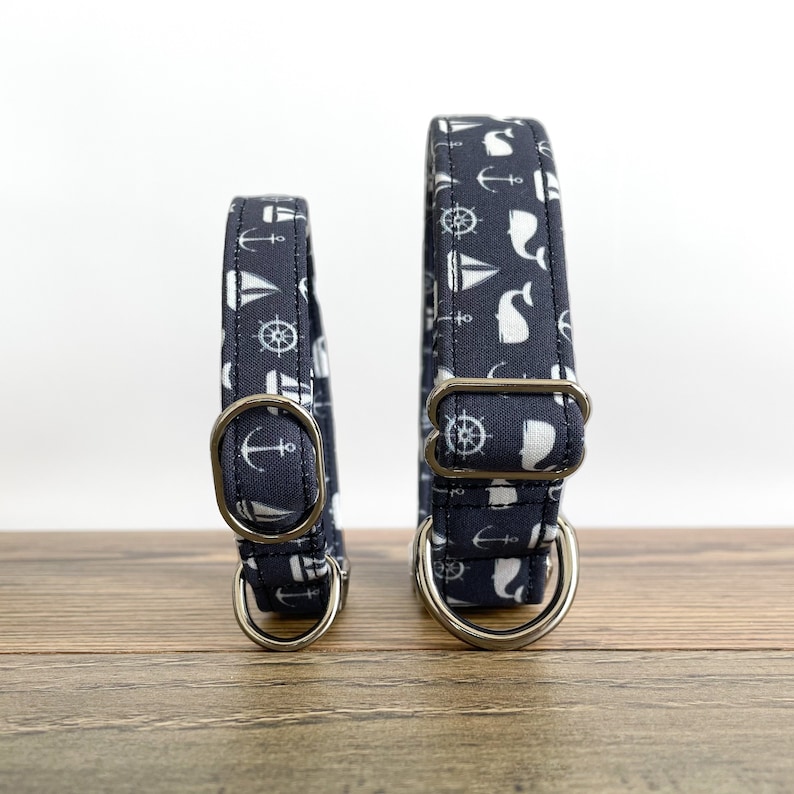 May include: Two navy blue dog collars with white nautical themed prints. The collars have silver metal D-rings. The smaller collar is on the left and the larger collar is on the right.