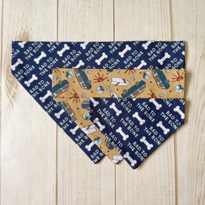 May include: A triangular pet bandana featuring a dark blue fabric with a repeating white pattern of dog bones and the text "BAD TO THE BONE" alongside a tan fabric section decorated with a colorful print of camper vans, suns, and seashells.