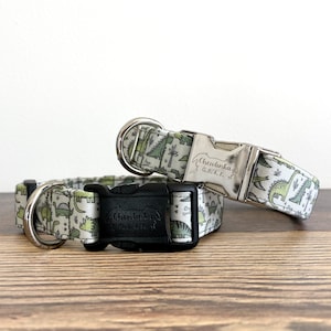 May include: Two dog collars with a dinosaur print. The collars are both white with green dinosaurs and have silver and black buckles. The silver buckle has the text "Chewlinka G.e.k.k." engraved on it.