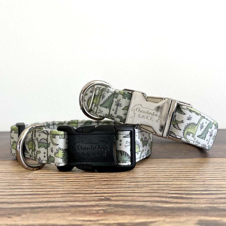 May include: Two dog collars with a dinosaur print. The collars are both white with green dinosaurs and have silver and black buckles. The silver buckle has the text "Chewlinka G.e.k.k." engraved on it.