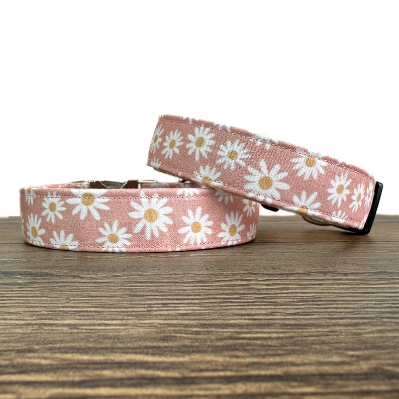 May include: Two pink dog collars with a white daisy print. The collars are made of fabric and have a silver buckle.