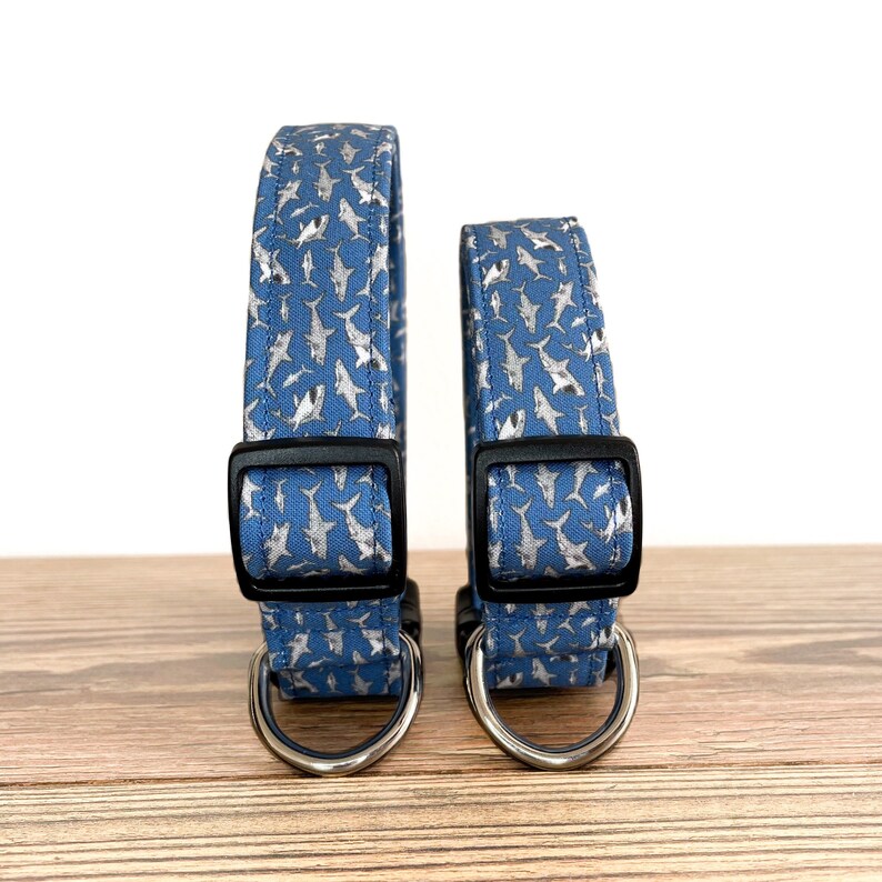 May include: Two dog collars with a blue background and a silver shark pattern. The collars have black buckles and silver D-rings.