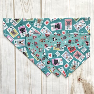 May include: A turquoise dog bandana with a repeating pattern of dogs wearing different accessories and hats. The bandana is folded in thirds, showing the different layers of the pattern.