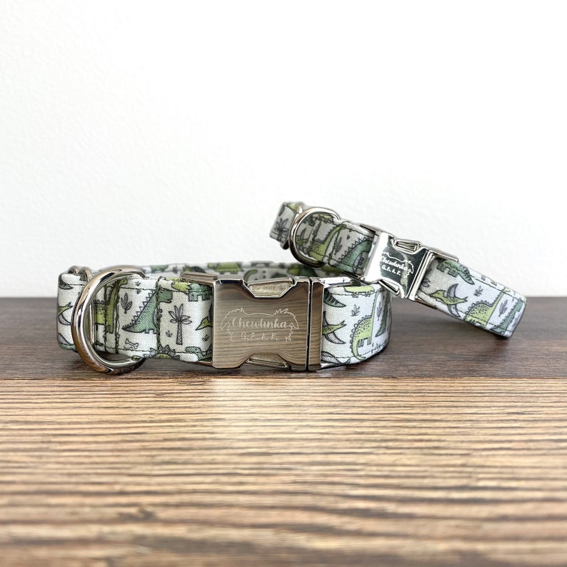 May include: Two dog collars with a green and white dinosaur print. The larger collar has a silver buckle with the text "Chewtinka's" engraved on it.