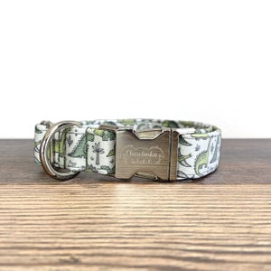 May include: A white dog collar with a green dinosaur print and a silver buckle. The buckle is engraved with "Chewlinka Gear."