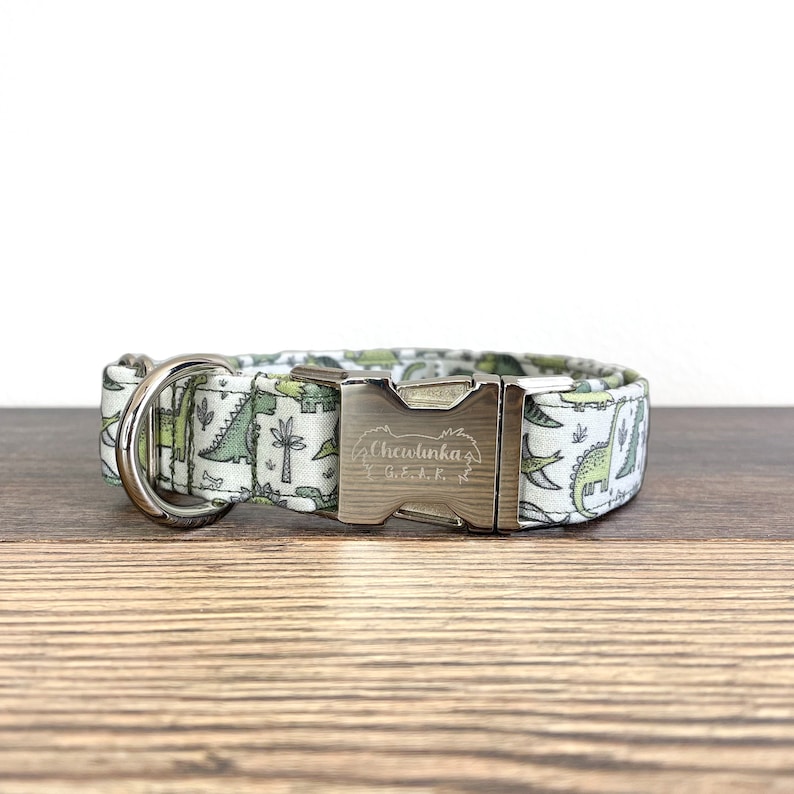 May include: A white dog collar with a green dinosaur print and a silver buckle. The buckle is engraved with "Chewlinka Gear."