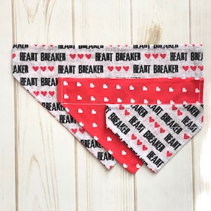 May include: Two dog bandanas with a red and white heart pattern. The top bandana has the text "HEART BREAKER" repeated in black. The bottom bandana has the text "HEART BREAKER" repeated in black on a white background.