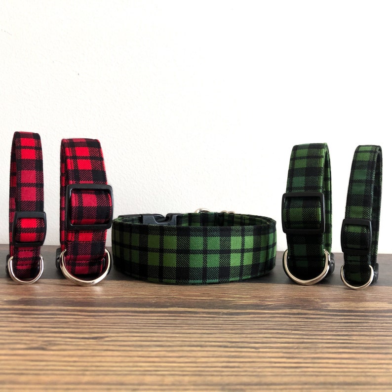 May include: Three dog collars in red and green plaid patterns. The collars are made of fabric and have black plastic buckles. The collars are arranged in a row on a wooden surface.