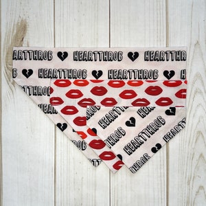 May include: A pink and white dog bandana with a black and white print of the words "Heartthrob" and a broken heart. The bandana also has a red lip print.