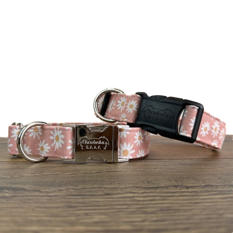 May include: Two pink dog collars with white daisy print. The collars have silver buckles with the text "Chewlunka G.E.A.R." engraved on them. One collar has a black buckle.