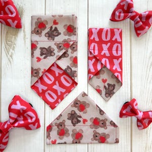 May include: Three dog bandanas with a Valentine's Day theme. One bandana features a pink and red pattern with the text "XOXO". Another bandana features a beige background with brown teddy bears and red hearts. The third bandana features a beige background with brown teddy bears and red hearts.  Two red bow ties with the text "XOXO" are also shown.