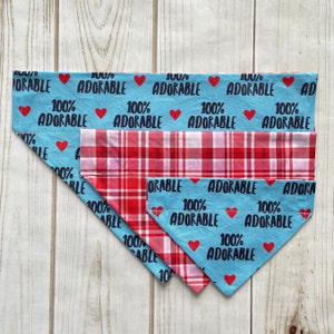 May include: Three dog bandanas in different sizes. The largest bandana is light blue with white hearts and the text "100% Adorable". The medium bandana is red and white plaid. The smallest bandana is light blue with white hearts and the text "100% Adorable".