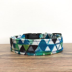 May include: A white dog collar with a geometric pattern in shades of blue, green, and black. The collar has a black plastic buckle.