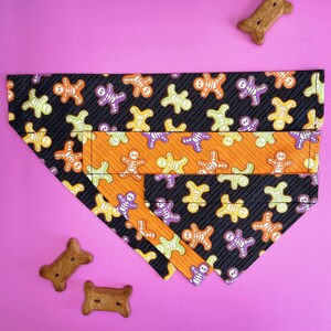 May include: A black, orange, and purple dog bandana with a skeleton pattern. The bandana is folded in a triangle shape and has a layer of orange fabric with a skeleton pattern on top of the black fabric.