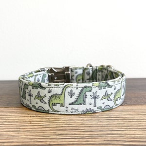 May include: A white dog collar with a green dinosaur print. The collar has a silver buckle and is made of fabric.