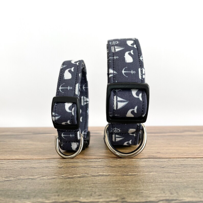 May include: Two dog collars with a nautical theme. The collars are navy blue with white anchors, sailboats, and whales. The collars are made of fabric and have a black buckle and a silver D-ring.