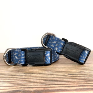 May include: Two blue dog collars with a shark print and silver buckles. The collars are made by "The Handmade Pet Co."