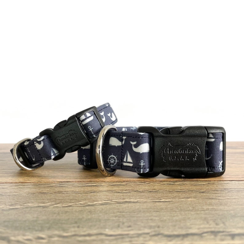 May include: Two navy blue dog collars with white whale, anchor, and sailboat designs. The collars have black plastic buckles and silver metal D-rings. The larger collar has the text "Chesapeake Bay Gift" on the buckle.