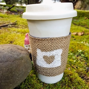 May include: A white to-go coffee cup with a burlap sleeve. The sleeve has a white outline of the state of Oregon with a heart in the middle.