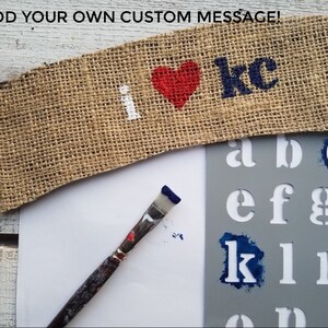 May include: A burlap banner with the words "I love kc" in white paint and a red heart. A stencil with letters of the alphabet is shown with blue paint on the letters "c" and "k". A paintbrush is also shown.
