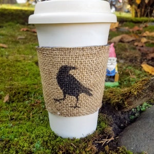 Peut inclure: Un gobelet à café blanc à emporter avec une gaine en toile de jute représentant une silhouette de corbeau noir. La gaine a un bord effiloché et est enroulée autour du gobelet.