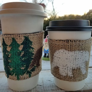 Pode incluir: Duas xícaras de café de papel branco com tampas, cada uma envolta em uma manga de serapilheira. Uma manga apresenta pinheiros verdes, a outra uma silhueta de urso branco. As xícaras estão em uma superfície de madeira.