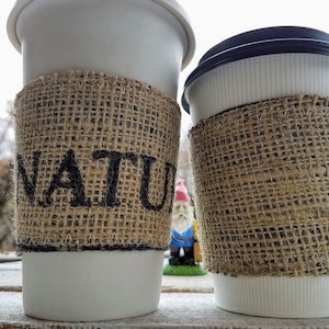 May include: Two white takeaway coffee cups with brown hessian sleeves. The sleeve on the left cup has the word "NATURE" printed on it in black.