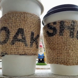 Op de afbeelding: Twee witte koffiebekers om mee te nemen met bruine jutehoezen. De hoes op de linker beker heeft het woord "OAK" erop gedrukt. De hoes op de rechter beker heeft het woord "STE" erop gedrukt.