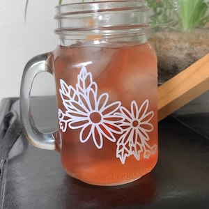May include: A clear glass mason jar mug with a white floral design. The mug is filled with a pink iced drink.