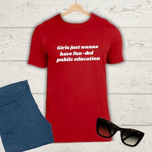 May include: A red t-shirt with the slogan "Girls just wanna have fun-ded public education" in white. The shirt is shown with blue jeans and tortoiseshell sunglasses.