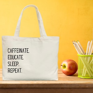 May include: White canvas tote bag with the text "CAFFEINE. EDUCATE. SLEEP. REPEAT." printed in black. The bag is sitting on a wooden surface with a red apple and a green pencil holder with pencils.