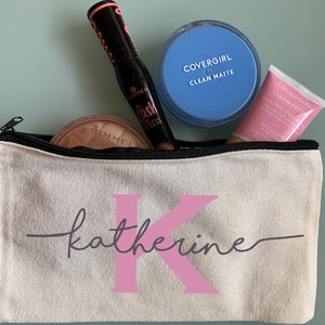 Peut inclure: Une pochette zippée en toile blanche avec le nom "katherine" écrit en cursive avec une initiale "K" rose. La pochette est remplie de produits de maquillage, dont un compact bleu, un tube rose et un mascara noir.