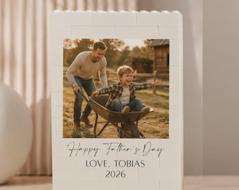 May include: A white rectangular block frame constructed from interlocking plastic bricks featuring a centred photograph of an adult pushing a child in a wheelbarrow. Below the image, black text reads Happy Father's Day, LOVE, TOBIAS, 2026.