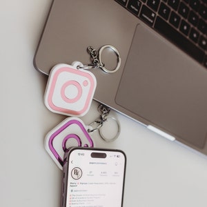 May include: Two pink and purple Instagram logo keychains with silver key rings on a laptop. The laptop screen shows a phone with the Instagram profile of papercutznlabels. The profile text reads "Sherry Signage | Laser Keychains | Apparel" and "The Creator of Smart Business Solutions, DIY of Grandkids' School Projects, Events & Exclusive Custom Signage, Seating Charts, papercutz.com, cutting and more".