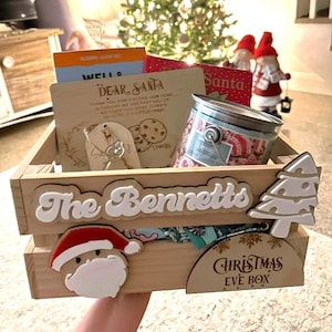 May include: A wooden Christmas Eve box with the name "The Bennetts" on the front. The box is filled with treats, including cookies, a key, and a candle. The box also has a Santa Claus face, a Christmas tree, and a sign that says "Christmas Eve Box".
