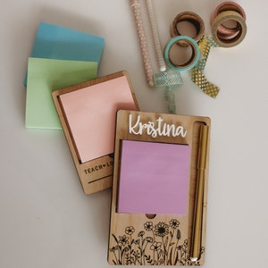 May include: A wooden desk organiser with the name "Kristina" engraved on it. It has a space for sticky notes and a gold pen holder. The organiser has a floral design at the bottom and the words "Teach Love" are engraved on the top.