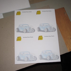 Peut inclure: Quatre feuilles de papier imprimées avec une illustration d'une voiture de course bleue et le texte "The Body Shop Omaha" répété quatre fois.