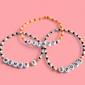 Op de afbeelding: Drie armbanden met kralen met witte letterkralen die "WEMBY 1", "ASTROS" en "SPURS" spellen. De armbanden hebben een mix van zwarte, witte, oranje, gele en blauwe kralen. De achtergrond is effen roze.