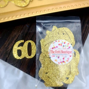 May include: Gold glitter confetti in a clear plastic bag with the text "The Fetti Boutique Balloons and Party Decor". There is a large gold glitter number 60 on a dark wood background.