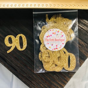 Op de afbeelding: Gouden glitter papieren uitsparingen van het nummer 90 en het woord "Negentig" in een doorzichtige plastic zak. De zak heeft een sticker met de tekst "The Fetti Boutique Balloons and Party Decor". Op een donker houten oppervlak.