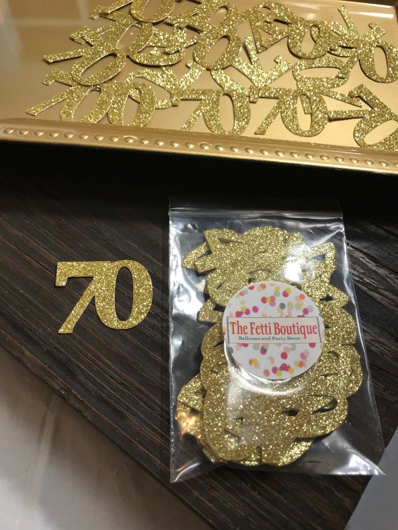 以下が含まれることがあります： 透明なプラスチック袋に入っている金色のグリッターの紙吹雪。袋には「The Fetti Boutique Balloons and Party Decor」の文字が書かれている。木製の表面に大きな金色のグリッターの数字70が置かれている。