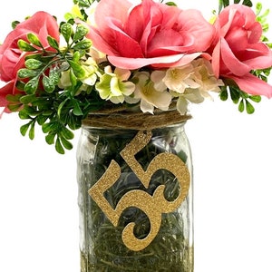 May include: A mason jar filled with green grass and pink flowers, decorated with a gold glitter number "55" for a birthday celebration.