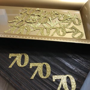 以下が含まれることがあります： 金色のトレイに金色のグリッターでできた数字70の切り抜き。暗い木製の表面に、より大きな金色のグリッターでできた数字70の切り抜きが3つある。