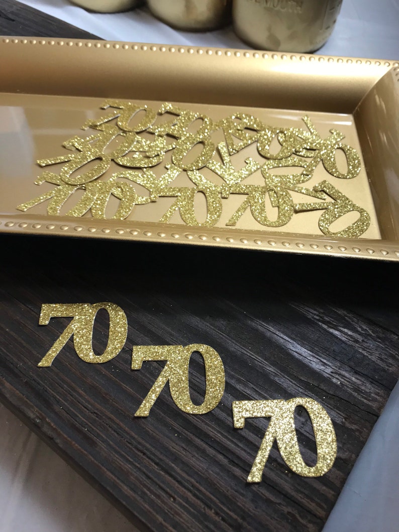 以下が含まれることがあります： 金色のトレイに金色のグリッターでできた数字70の切り抜き。暗い木製の表面に、より大きな金色のグリッターでできた数字70の切り抜きが3つある。