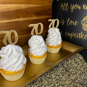 May include: Three cupcakes with white frosting and gold glitter "70" toppers. The cupcakes are on a gold tray with a gold glitter tablecloth. A black sign with gold lettering reads "All you need is love and cupcakes."