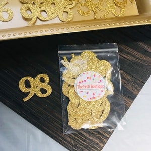 May include: Gold glitter confetti in a clear plastic bag with a white label that says "The Fetti Boutique" with a colorful confetti design. The bag is on a dark brown wooden surface. There are also gold glitter numbers on the surface.